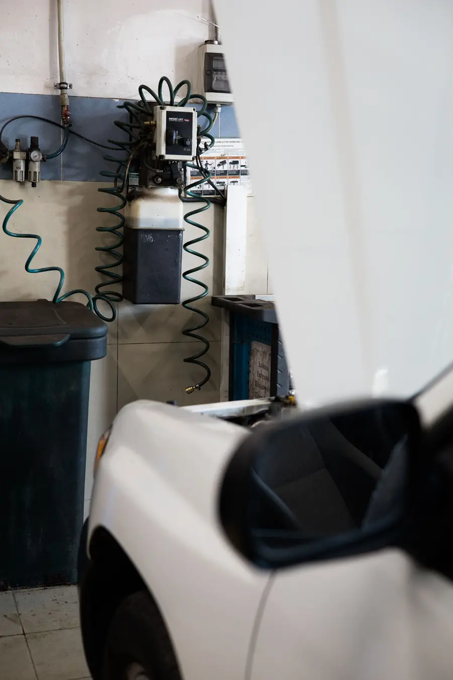The front corner of a white car parked inside a repair shop, with air lines on the wall